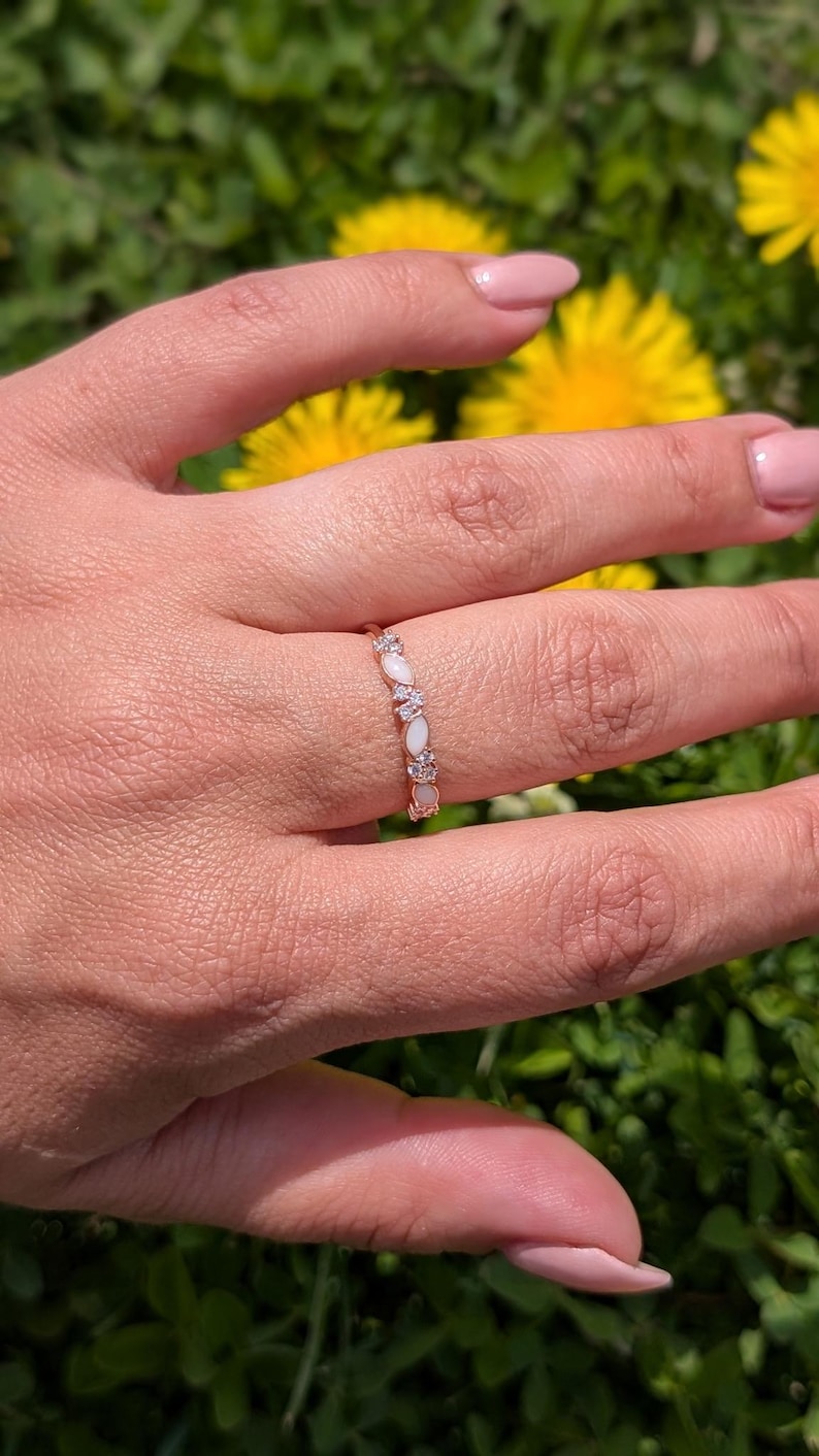 May include: A gold-toned ring with alternating oval-shaped white gemstones and small clear stones. The ring is worn on a hand with pink nail polish, set against a backdrop of green grass and yellow flowers.