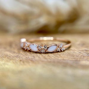 May include: Rose gold ring featuring a series of white, marquise-shaped stones interspersed with clusters of small, clear gemstones. The ring is set on a wooden surface, showcasing its delicate design and sparkle.