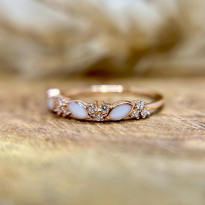 May include: Rose gold ring featuring a series of white, marquise-shaped stones interspersed with clusters of small, clear gemstones. The ring is set on a wooden surface, showcasing its delicate design and sparkle.