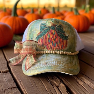 Könnte beinhalten: Eine grüne und weiße Trucker-Mütze mit einem Kürbis-Design. Die Mütze hat eine farbenfrohe Kürbisgrafik mit einer grünen Schleife, eine Holzperlenverzierung und ein kariertes Band. Die Mütze ist vor einem Hintergrund aus Kürbissen platziert.