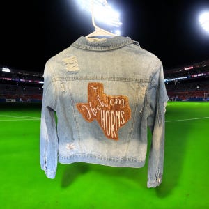 Könnte beinhalten: Hellblaue Jeansjacke mit Distressed-Details. Verfügt über einen braunen und weißen Aufnäher in Form von Texas mit den Worten "Hook 'em Horns". Die Jacke hängt an einem weißen Kleiderbügel.