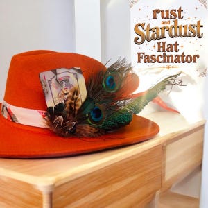 May include: A bright orange fedora hat with a white ribbon and a peacock feather accent. The hat also features a vintage playing card tucked into the band. The text "rust and stardust Hat Fascinator" is visible in the background.