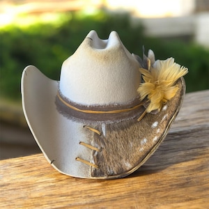 May include: A cream-colored cowboy hat with a two-tone design. The hat features a light-colored crown and brim, with a section of patterned fur. It is adorned with a brown band, yellow stitching, and a cluster of yellow feathers.