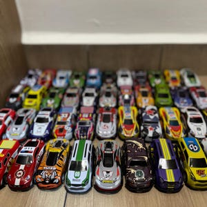 Puede incluir: Una colección de coches de carreras de juguete en varios colores y diseños. Los coches presentan gráficos detallados, incluyendo rayas de carreras, logotipos de patrocinadores y números. El surtido incluye coches rojos, amarillos, verdes, azules y blancos, dispuestos en filas.