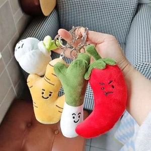 May include: A selection of plush vegetable keyrings, featuring a white radish, yellow ginger, green broccoli, and a red chilli pepper. Each keyring has a smiling or angry face and is attached to a silver keyring.