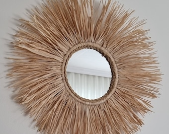 May include: A round mirror with a natural woven straw frame. The frame is thick and has a sunburst design. The mirror is reflecting a white wall.