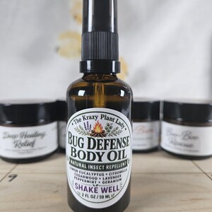 May include: A brown glass spray bottle of The Krazy Plant Lady Bug Defense Body Oil, a natural insect repellent. The white label features a campfire graphic and lists ingredients including lemon, eucalyptus, citronella, cedarwood, lavender, peppermint, and geranium. The bottle contains 2 fl oz.