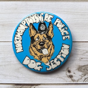 May include: A blue and white button with a cartoon German Shepherd wearing a police hat. The text around the dog reads "Northamptonshire Police Dog Section".