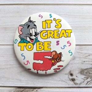 May include: A white button featuring a cartoon image of Tom and Jerry. The button reads "It's Great To Be 5" with the number 5 in red and surrounded by the number 5 in various colours.