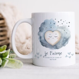 Puede incluir: Taza de cerámica blanca con un diseño de acuarela azul de una nube con estrellas y un corazón con una cara. El texto "je t'aime jusqu'à la lune et retour" está impreso en la taza.