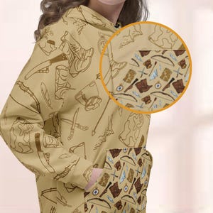 May include: Beige hoodie featuring a repeating pattern of brown adventure-themed illustrations. The design includes bows, arrows, maps, compasses, and boots. The hoodie has a kangaroo pocket with a smaller, detailed version of the pattern.