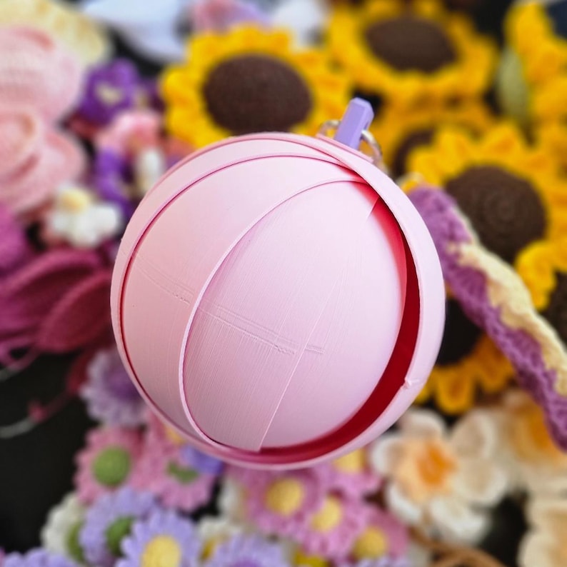 May include: A pale pink spherical ornament with curved, overlapping sections, similar to a flower bud. It has a small purple loop at the top. The background shows various colourful crocheted flowers, including sunflowers.