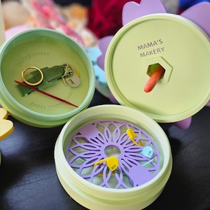 May include: Three green round containers with lids, each with a different design. The first container has a green felt pad, a gold ring, a red needle, and a small silver tool. The second container has a purple flower design and a small orange tool. The third container has a purple flower design with a green outline. The lids of the containers have the text "Mama's Makery" printed on them.