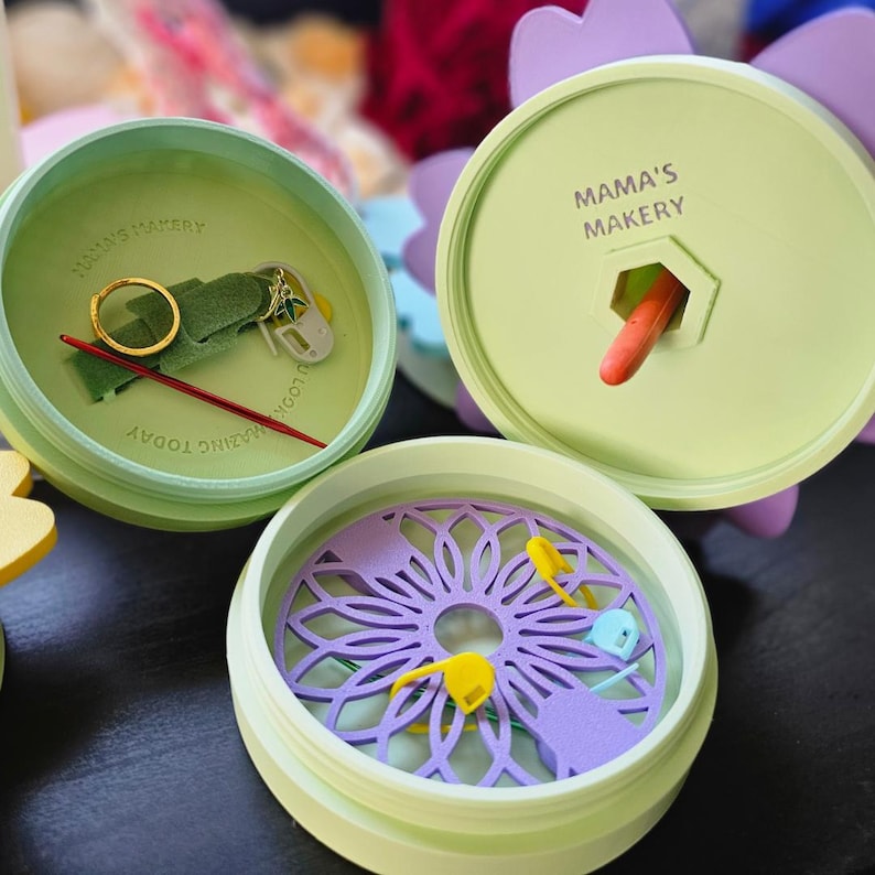 May include: Three green round containers with lids, each with a different design. The first container has a green felt pad, a gold ring, a red needle, and a small silver tool. The second container has a purple flower design and a small orange tool. The third container has a purple flower design with a green outline. The lids of the containers have the text "Mama's Makery" printed on them.