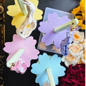 May include: Five wooden flower-shaped stands with pastel colors, each with a crocheted item hanging from a metal ring. The stands are arranged on a dark surface.