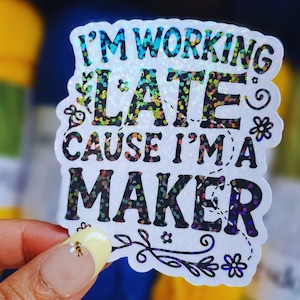 May include: A white sticker with a glittery rainbow finish. The sticker says "I'm working late cause I'm a maker" with floral designs around the text.