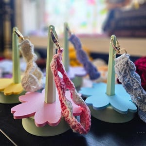 May include: Three crocheted straps displayed on pastel-colored flower-shaped stands. The stands are made of foam and have a light green base. The straps are in pink, blue, and white.