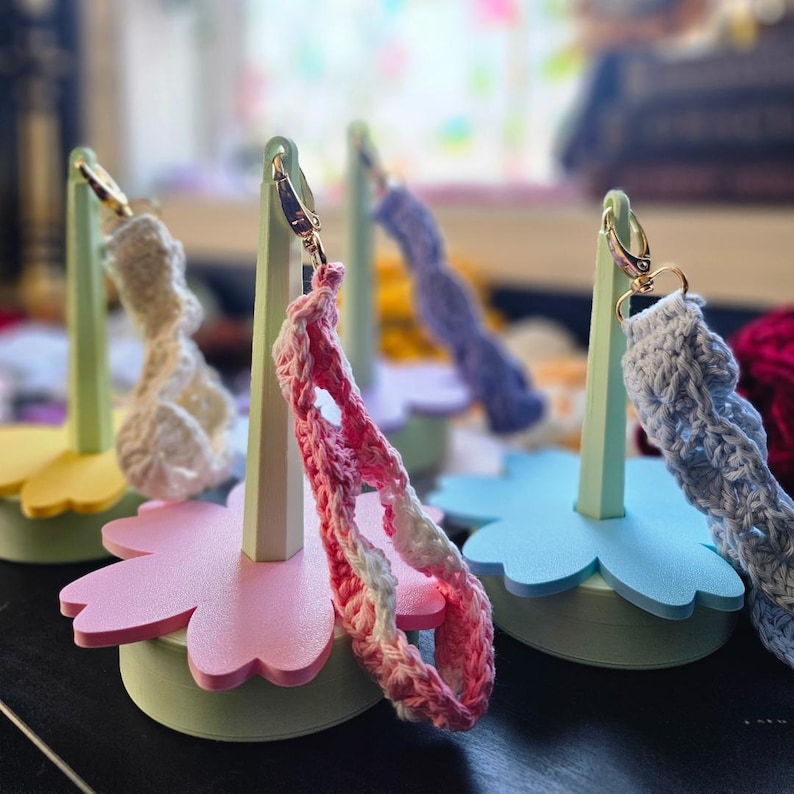 May include: Three crocheted straps displayed on pastel-colored flower-shaped stands. The stands are made of foam and have a light green base. The straps are in pink, blue, and white.