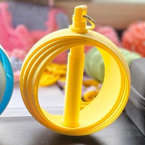May include: A yellow yarn swift, used for winding yarn into a ball. The swift features a circular design with multiple rings and a central post. A small metal ring is attached to the top. Other yarn colours are visible in the background.