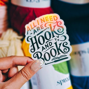 May include: A holographic sticker with the text "ALL I NEED ARE HOOKS AND BOOKS" in black and orange. The sticker is held in a hand, with yarn in the background.