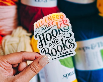 May include: A holographic sticker with the text "ALL I NEED ARE HOOKS AND BOOKS" in black and orange. The sticker is held in a hand, with yarn in the background.