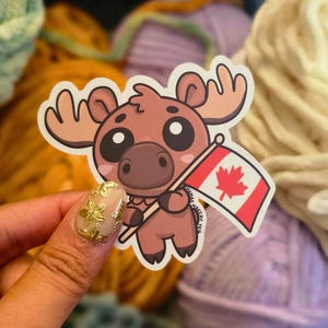 May include: A brown cartoon moose holding a Canadian flag. The moose has large eyes and a friendly expression. The flag is red with a white maple leaf.