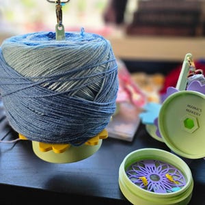 May include: A blue and white yarn ball is on a green and yellow yarn bowl. The yarn bowl has a flower design. A green and purple yarn bowl with a flower design is in the background. The yarn bowl has the text "Mama's Makery" on it.