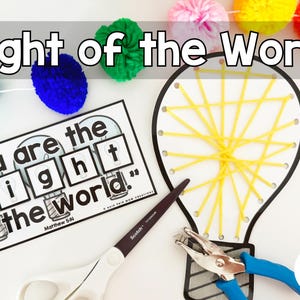Op de afbeelding: Een knutselproject met de tekst "Light of the World". Het bevat een gloeilampvormige draadkunst, een papier met de tekst "You are the light of the world", een schaar en een perforator. Kleurrijke pompons zijn bovenaan geregen.