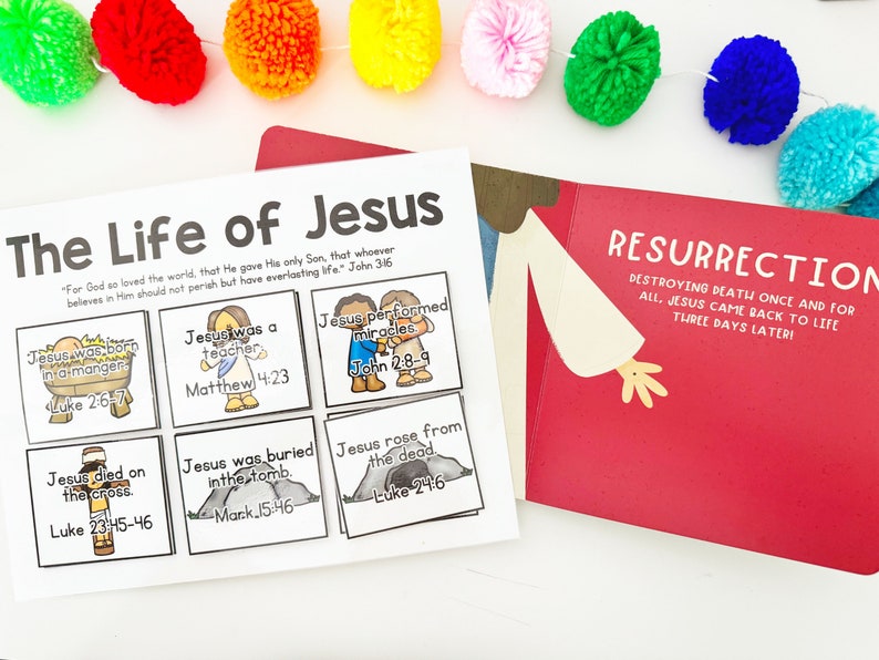May include: A children's book and a printed page with illustrations about the life of Jesus. The book is red with the word "Resurrection" and the printed page has the title "The Life of Jesus". Colorful pom-poms are strung across the top.