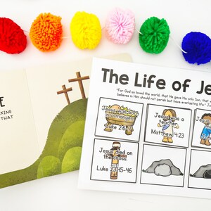 May include: A colorful pom-pom garland hangs above an open book and a printed sheet titled "The Life of Jesus." The book shows a hillside with three crosses. The sheet has illustrated scenes from Jesus' life with Bible verses.