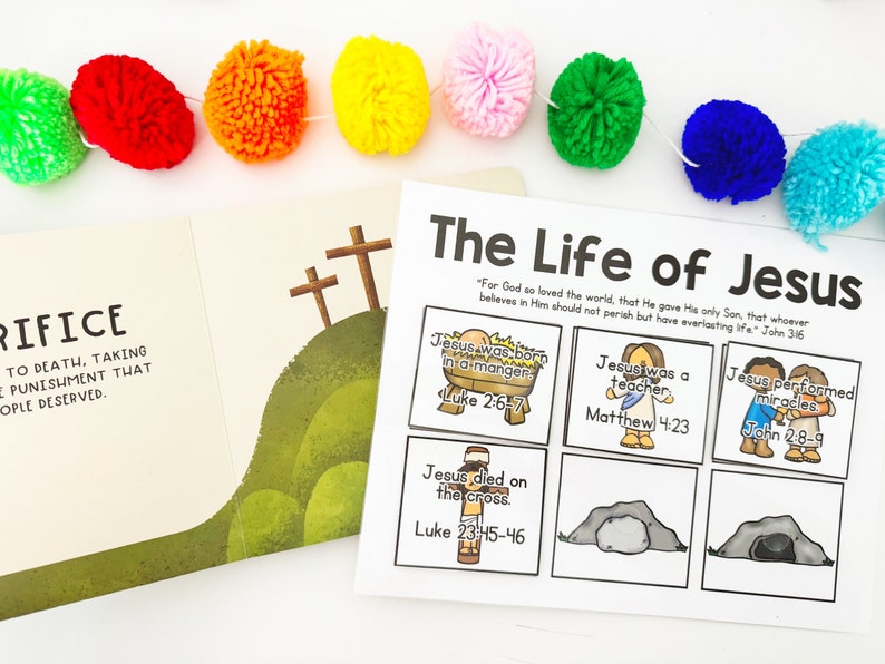 May include: A colorful pom-pom garland hangs above an open book and a printed sheet titled "The Life of Jesus." The book shows a hillside with three crosses. The sheet has illustrated scenes from Jesus' life with Bible verses.