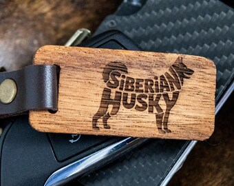 May include: Wooden keychain with a brown leather strap and a metal clasp. The rectangular keychain features a laser-engraved silhouette of a Siberian Husky with the words "Siberian Husky" within the dog's outline. The keychain rests on a car key and a carbon fiber wallet.