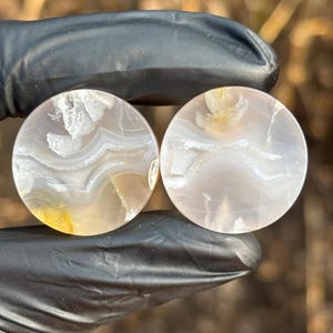 May include: Two circular, polished stone plugs featuring a translucent, pale lavender and white banded agate pattern. The stones display natural, swirling inclusions and soft, milky white layers throughout their smooth, rounded surfaces.