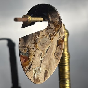 May include: A distinctive, teardrop-shaped earring plug with a natural, marbled pattern in beige, brown, and black. The plug is held by a brass rod, highlighting its intricate details and organic texture. The earring is a statement piece.