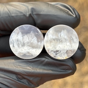 May include: A pair of circular ear plugs made of translucent, light gray stone. Each plug features a distinct, natural white plume-like inclusion pattern that resembles delicate, frost-covered foliage suspended within the smooth, polished surface.