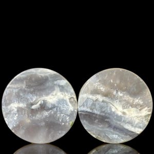 Puede incluir: Dos piedras redondas y pulidas con aspecto veteado. Las piedras presentan una mezcla de áreas grises, blancas y translúcidas, creando un patrón nublado y natural. Las piedras están sobre un fondo negro.