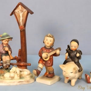 Puede incluir: Colección de figuras de porcelana vintage. Las figuras representan niños tocando instrumentos musicales, un niño montado en un cerdo y un ángel. También se incluye una cruz de madera con una figura religiosa. Las figuras están pintadas en varios colores.