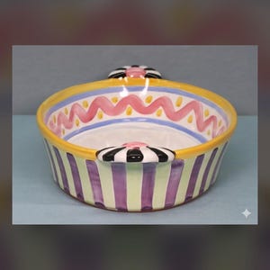 May include: A round ceramic bowl featuring a yellow rim, a pink wavy line pattern on the interior, and vertical purple and light green stripes on the exterior. The bowl includes two handles decorated with black and white stripes and a pink center.