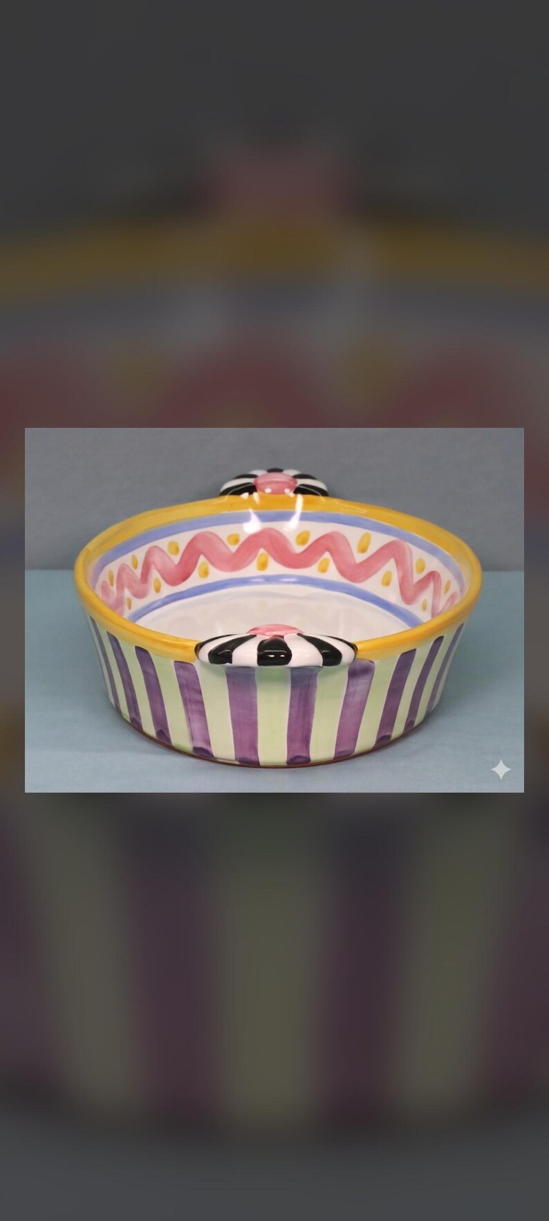 May include: A round ceramic bowl featuring a yellow rim, a pink wavy line pattern on the interior, and vertical purple and light green stripes on the exterior. The bowl includes two handles decorated with black and white stripes and a pink center.