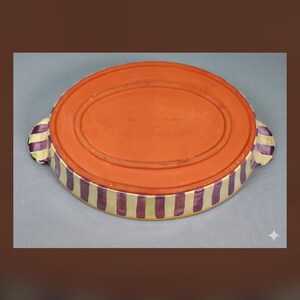 May include: An oval ceramic baking dish with a terracotta-colored base and sides featuring vertical purple and cream-colored stripes. The dish includes two small handles on opposite ends, each decorated with the same purple and cream striped pattern.