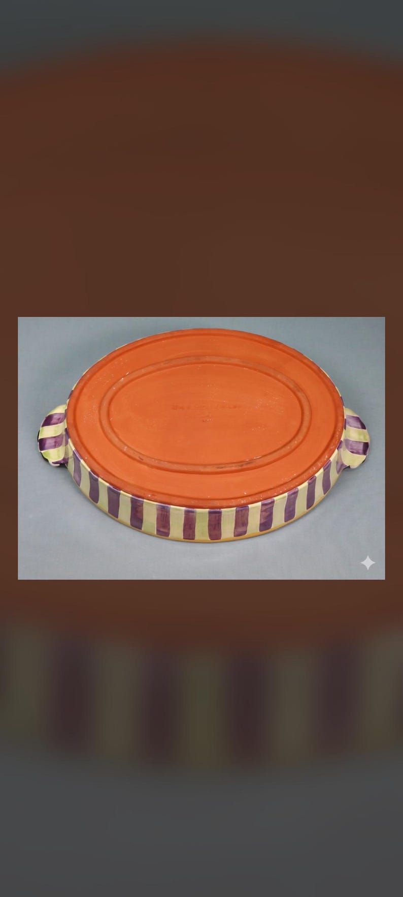 May include: An oval ceramic baking dish with a terracotta-colored base and sides featuring vertical purple and cream-colored stripes. The dish includes two small handles on opposite ends, each decorated with the same purple and cream striped pattern.