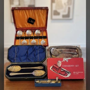 May include: A collection of vintage silver-plated serving utensils including a set of six shell-shaped spoons in a red case, two large ornate spoons in a dark blue case, and a Wm. Rogers Silver cranberry set with a tray and slotted spoon.