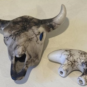 May include: A ceramic bull skull with a matching curved horn-shaped piece. The skull is off-white with dark, lightning-like patterns. The eye sockets are blue. The horn-shaped piece has similar patterns.
