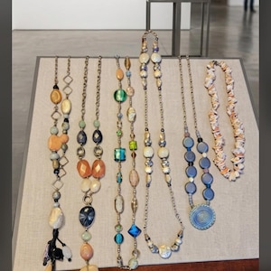 May include: Six necklaces displayed on a stand. The first features orange and black beads with a tassel. The second has orange and blue oval beads. The third contains green and blue geometric beads. The fourth has white and gold beads. The fifth has blue circular beads. The sixth is a shell necklace.