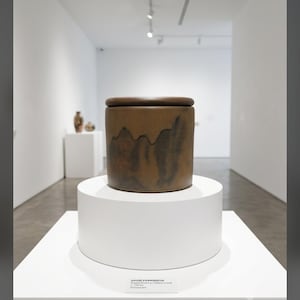 May include: A cylindrical lidded ceramic jar with a matte brown finish and dark, abstract vertical brushstroke patterns. The vessel features a flat, circular lid and sits on a white circular pedestal with a small white label displaying text.