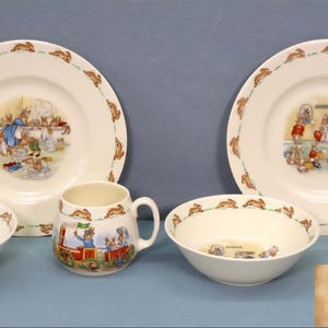 Könnte beinhalten: Ein Vintage-Geschirrset für Kinder mit Tellern, Schalen und Tassen. Die weißen Keramikteile haben einen braunen Hasenrand und farbenfrohe Illustrationen von Hasen. Das Set enthält zwei Teller, zwei Schalen und zwei Tassen.