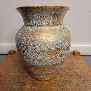 May include: A decorative vase with a wide base and flared rim. The vase has a gold metallic finish with a distressed, textured appearance. The body of the vase is a mix of gold and a light blue-green color.