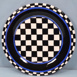 May include: A decorative ceramic plate with a black and white checkered center. The plate has a black border with a blue accent and a checkered pattern around the rim. The plate is round and has a glossy finish.