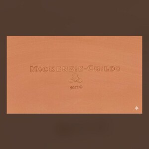 May include: A rectangular terracotta-colored surface featuring the embossed text MACKENZIE-CHILDS, a small floral emblem, and the year 2012. The surface has a smooth, matte finish with a consistent warm orange-brown tone throughout the entire area.