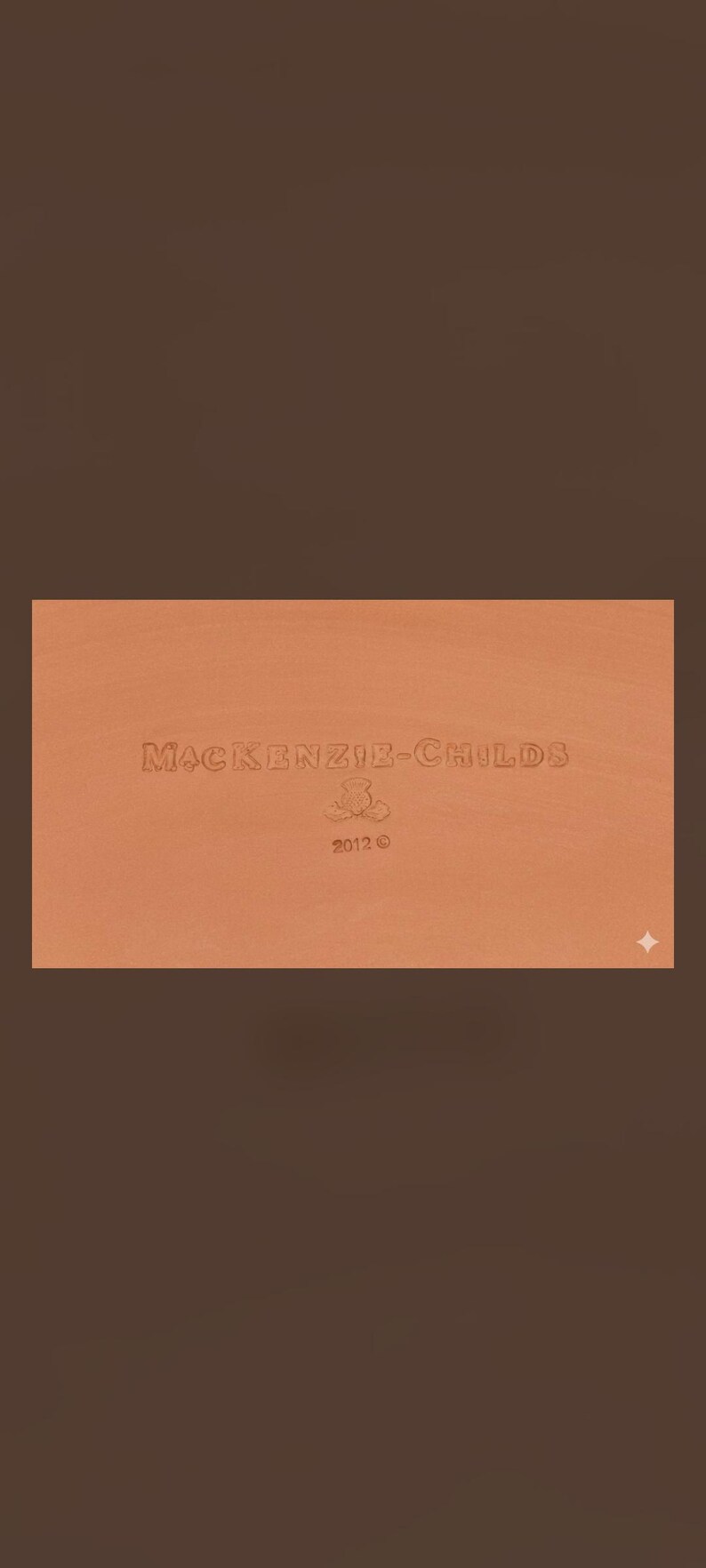 May include: A rectangular terracotta-colored surface featuring the embossed text MACKENZIE-CHILDS, a small floral emblem, and the year 2012. The surface has a smooth, matte finish with a consistent warm orange-brown tone throughout the entire area.